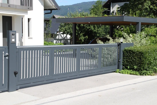 Scène extérieure résidentielle avec clôture en bois blanc à lames verticales espacées, piliers maçonnés beiges, végétation luxuriante en arrière-plan incluant arbres et arbustes verts