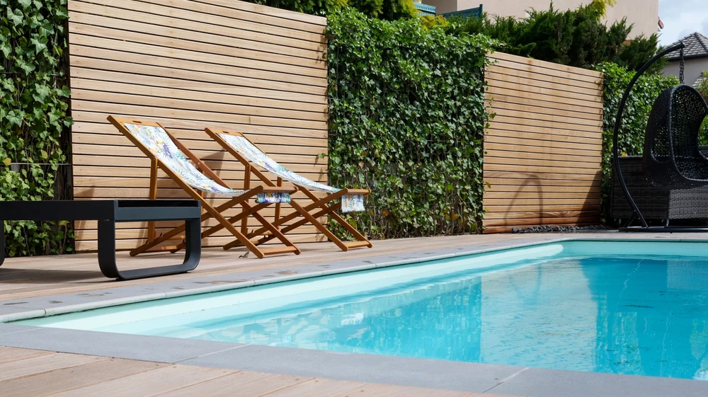 Photographie d'un jardin paysagé premium montrant un ensemble cohérent d'aménagements extérieurs : une terrasse pavée en pierre naturelle au premier plan, une clôture ou portail design en arrière-plan, des plantations variées et bien intégrées (arbustes, massifs fleuris, conifères), et des éléments de circulations aménagées (allées), tous harmonieuement composés dans une mise en scène globale d'aménagement extérieur complet et de haut standing